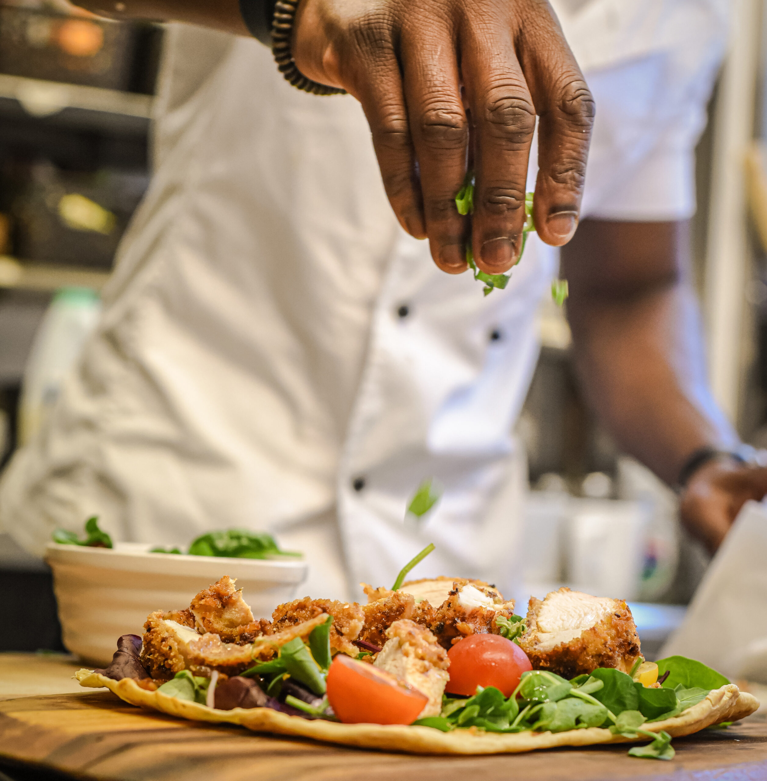 Chef sprinkles fresh green herbs over a crispy chicken flatbread topped with greens and tomatoes.